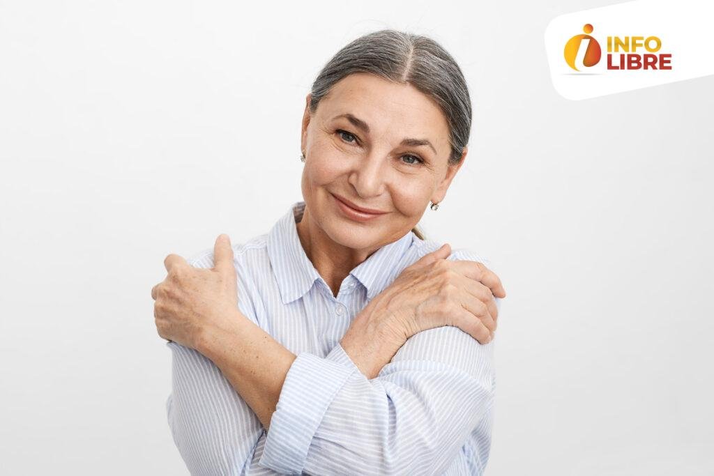 Horizontal shot of charming relaxed cheerful elderly senior female in blue shirt keeping hands on shoulders with arms on chest, hugging herself, looking at camera with joyful peaceful smile vejez